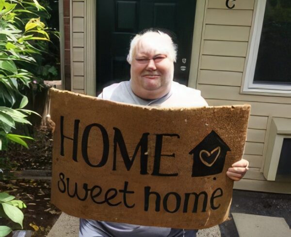 Steve holds a doormat that reads "home sweet home" outside his front door.
