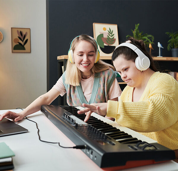 Two caucasian women wearing headphones playing together on an electronic keyboard. One of the women has downs syndrome, brown hair, and is wearing a yellow sweater. The other woman has blonde hair and is wearing a green and brown blouse.