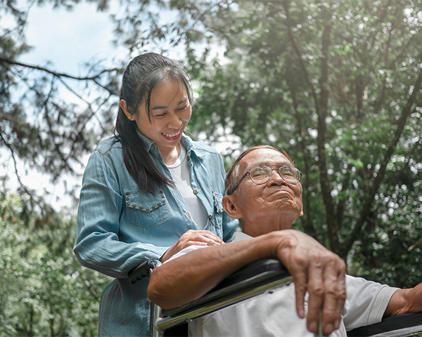 A young Asian woman wearing a denim shirt gently assists an elderly Asian man in a wheelchair outside in a sunny park. They are both smiling.