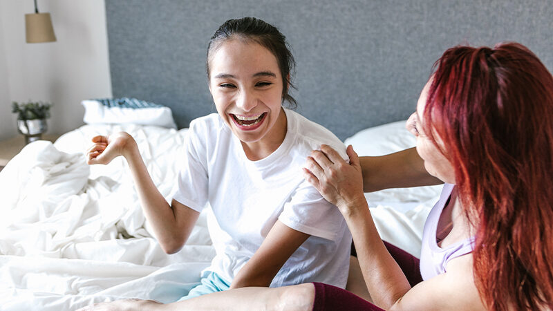 Two young women laugh together while seated on a bed, one with dark hair, and one with dyed red hair.