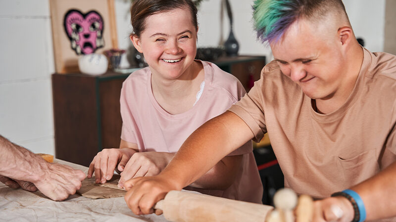Two Caucasian people with Downs syndrome smile together as they bake together.