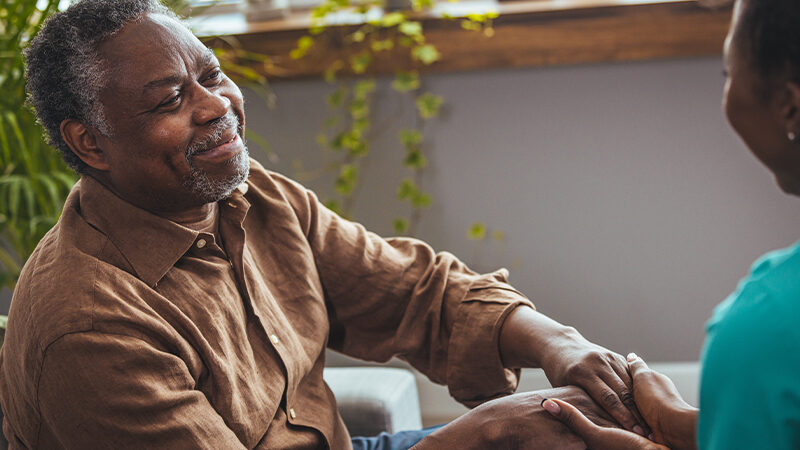 An elderly African-American man smiles as he gently holds the hands of an African-American woman, who smiles back at him.