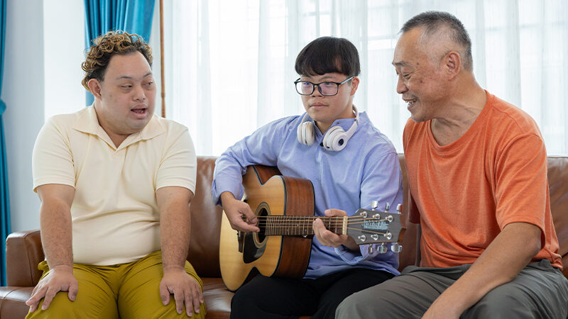 Two young Asian men with Downs syndrome sit beside an elderly Asian man on a couch in a living room. One of the younger men is playing an acoustic guitar. The left man is wearing a white shirt and yellow pants, the center man is wearing a blue button-down shirt and black pants, and the right man is wearing an orange shirt and grey pants.