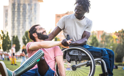 A Caucasian man and an African-American man sit in a park. The Caucasian man is wearing a pink shirt and is sitting down; he has a prosthetic leg. The African-American man is in a wheelchair. They are smiling at each other.