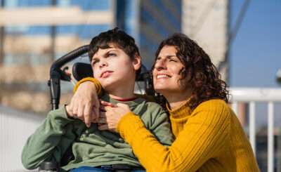 A Middle Eastern woman wearing a yellow sweater kneels beside a young Middle Eastern boy in a wheelchair. He is wearing a green shirt and they are outdoors, looking off in the distance.