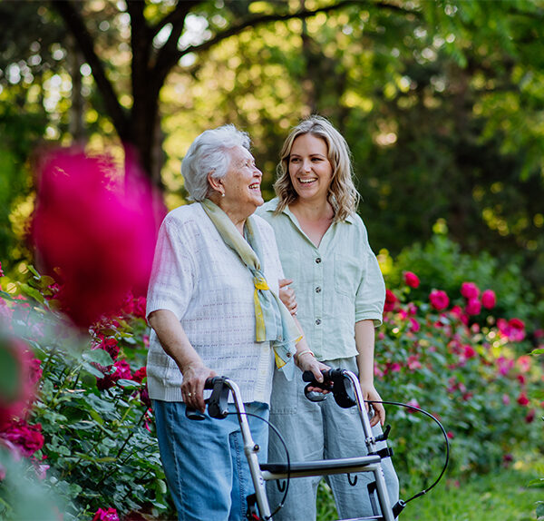 A Caucasian woman with blonde hair and a light green shirt walks beside an elderly Caucasian woman wearing a white shirt. They are in a rose garden.