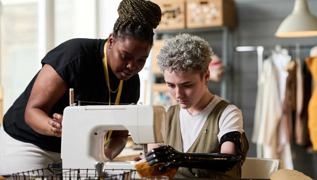 An African-American woman assists a Caucasian person with a sewing machine. The Caucasian person has a prosthetic arm.