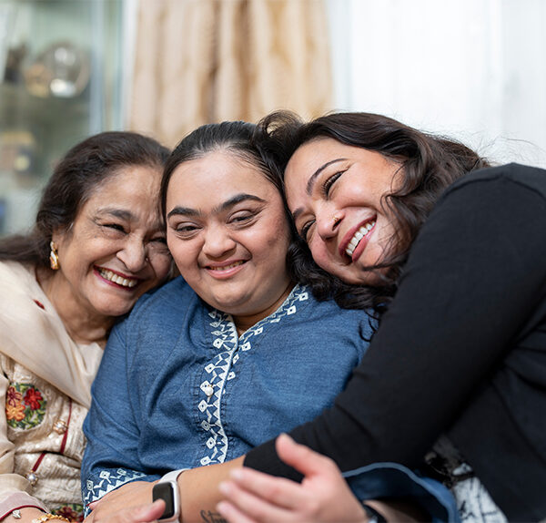 A trio of Middle Eastern women embrace and laugh together while seated on a couch in a living room. The left woman is elderly and wearing a beige outfit, the center woman has Downs syndrome and is wearing a blue outfit, and the right woman is middle-aged and wearing a black outfit.