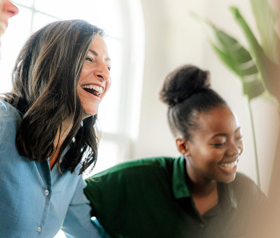 A Caucasian woman with dark hair, wearing a light blue shirt and an African-American woman with dark hair and a dark green shirt sit in a sun-filled room and are laughing.