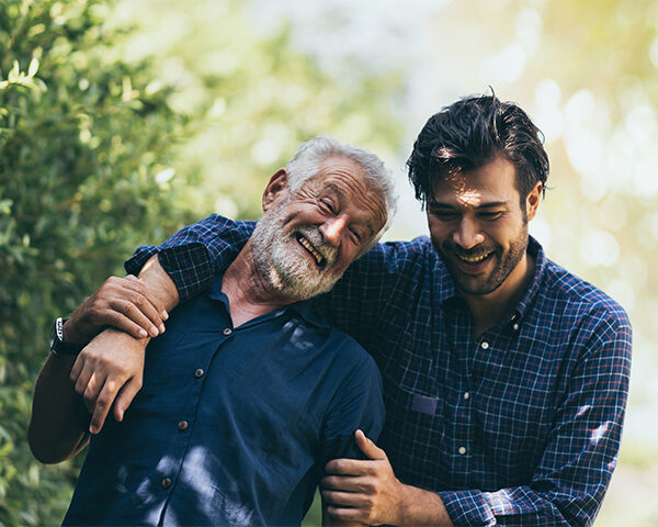 An older man with white hair and a beard and a younger man with black hair and a beard are walking in a park. Both men are wearing dark blue button-down shirts.