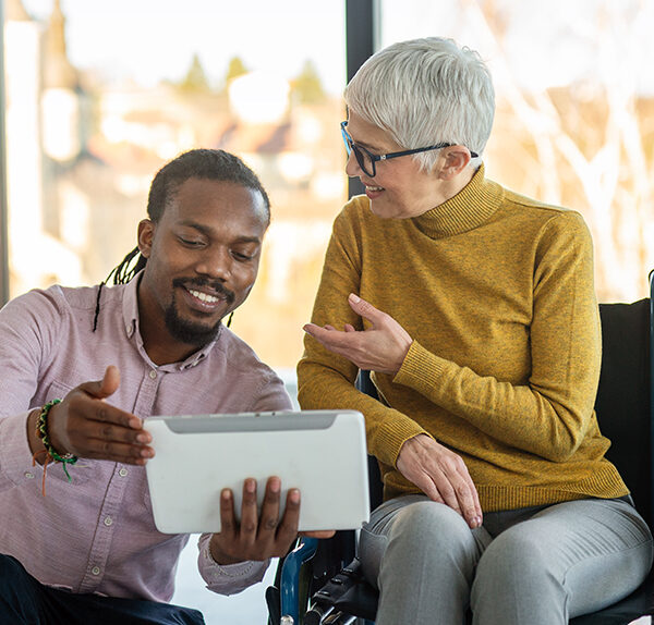 An African-American man wearing a pink button-down shirt shows an older Caucasian woman wearing a yellow turtleneck something on a digital tablet.