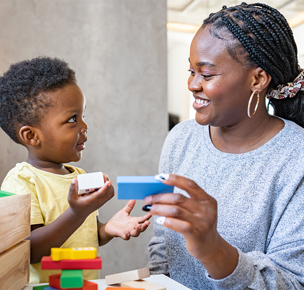 An African-American woman plays with an African-American toddler. They are both smiling, and the woman wears a light grey sweater and the boy wears a pale yellow shirt.