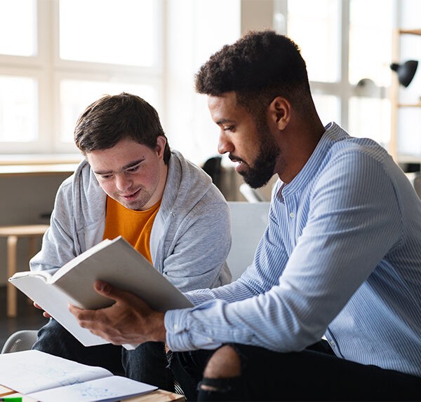 A Caucasian man with dark hair and Down syndrome wearing a light grey hoodie and orange shirt sits beside an African-American man with dark hair and a beard inside an office or apartment building. They are looking at a book together.