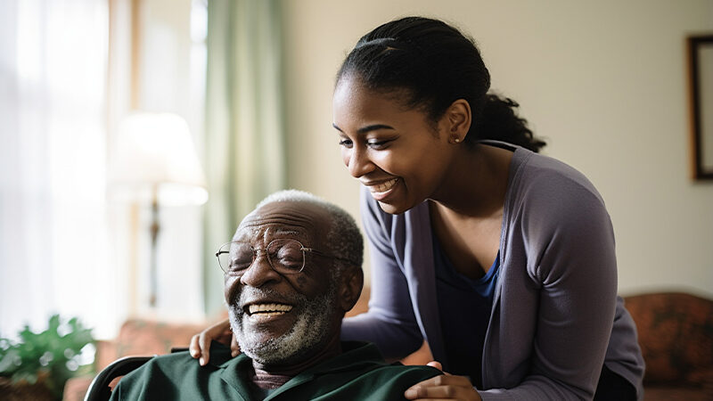 A young African-American girl with dark hair in a ponytail and wearing a lavender cardigan has her hands gently placed on the shoulders of an elderly African-American man with white hair and a beard, wearing a dark green shirt. They are both laughing and smiling and in a light-filled living room inside a house.