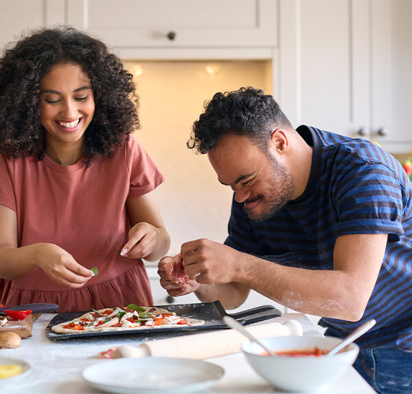 A young woman with dark curly hair and a red blouse makes a pizza with a young man with Downs syndrome and dark curly hair; he is wearing a blue shirt.