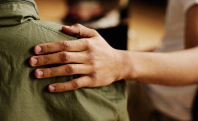 A woman of color places her hand compassionately on the shoulder of a person wearing a green jacket.