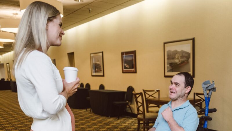 A blond woman, who is wearing a white sweater and an orange dress, is speaking to a man, wearing a blue shirt and grey pants, who is in a wheelchair.
