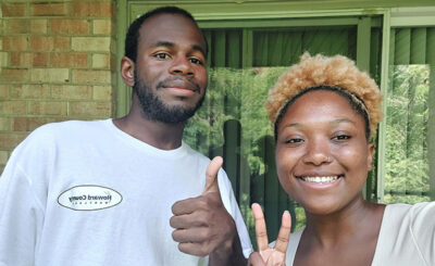 Both smiling at the camera, a young African-American male is giving a "thumbs up" sign next to an African-American woman who is holding her fingers in a "peace" sign.
