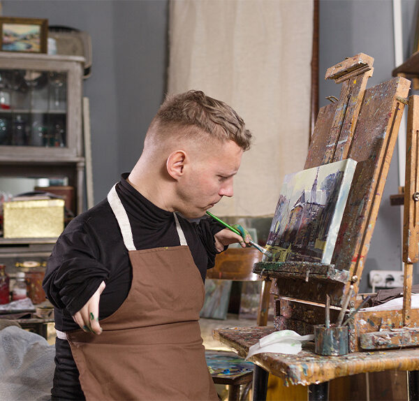 A Caucasian male with light hair and Dwarfism paints at an easel inside an artist studio. He is wearing a black shirt and a brown apron.