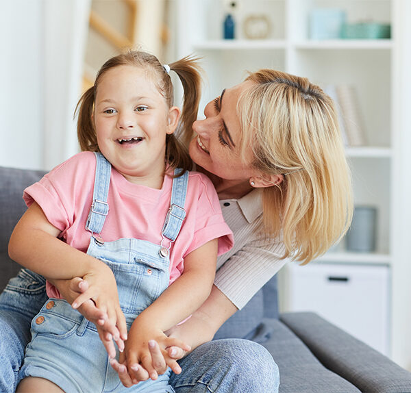 A blonde, Caucasian woman holds a child with Downs syndrome. They are both smiling and laughing. The child is wearing a pink shirt and denim overalls, and the woman is wearing an off-white shirt and denim jeans. They are in a clean, light-filled room inside a house.