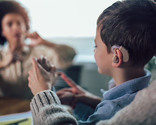 A young Caucasian boy with dark hair and a hearing aid is assisted by a person. A woman with dark curly hair sits across from the boy in the background, gesturing to him.