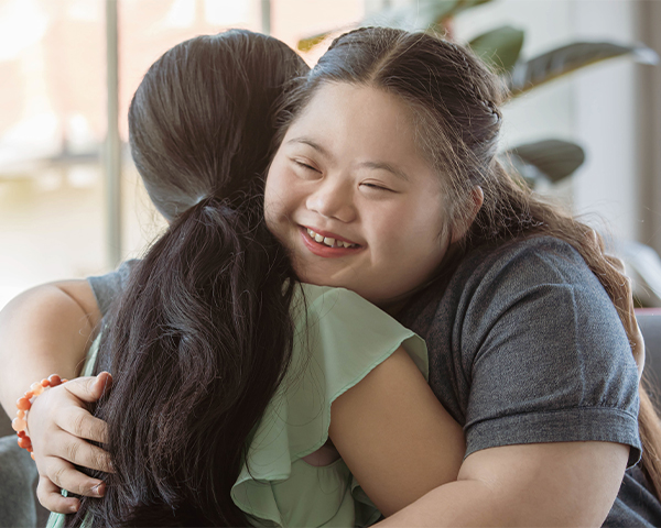 Two Asian women with dark hair embrace compassionately. One is facing away from the camera, the other is smiling. The smiling woman has Downs syndrome and is wearing a dark grey shirt. The other woman is wearing a light green blouse. They are inside a white, light-filled room within a house.