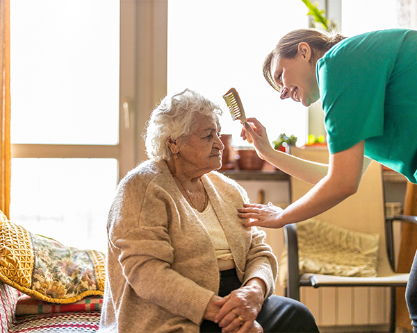 An elderly woman with curly white hair wearing a beige blouse and cardigan and dark pants is seated in a light-filled room. She is assisted by a smiling Caucasian woman wearing a bright green shirt. The younger woman is brushing the elderly woman’s hair.
