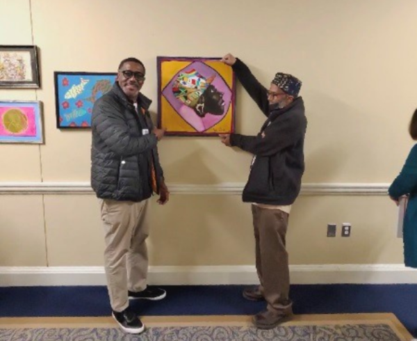 Image of Andre (right) display his artwork in an exhibit hall and his Employment Program Manager, Derrick Boyd from Shura, Inc. (left) posing with Andre.