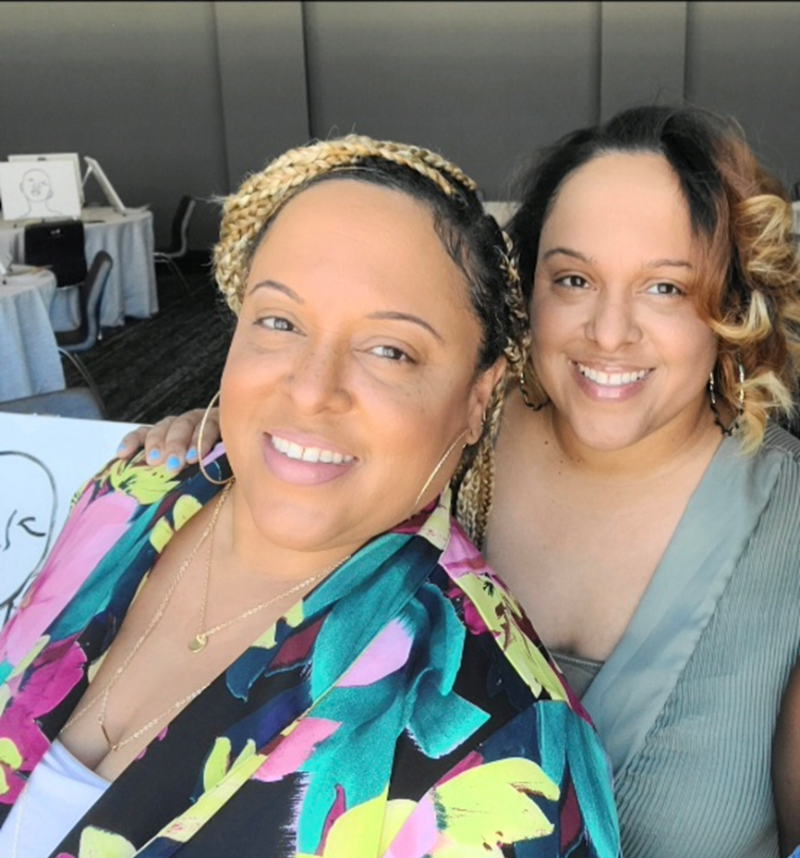 Two smiling twin sisters pose closely together indoors, one with blonde braided hair and a vibrant floral shirt, the other with curly dark hair with blonde tips and a muted green top, creating a warm, friendly atmosphere.
