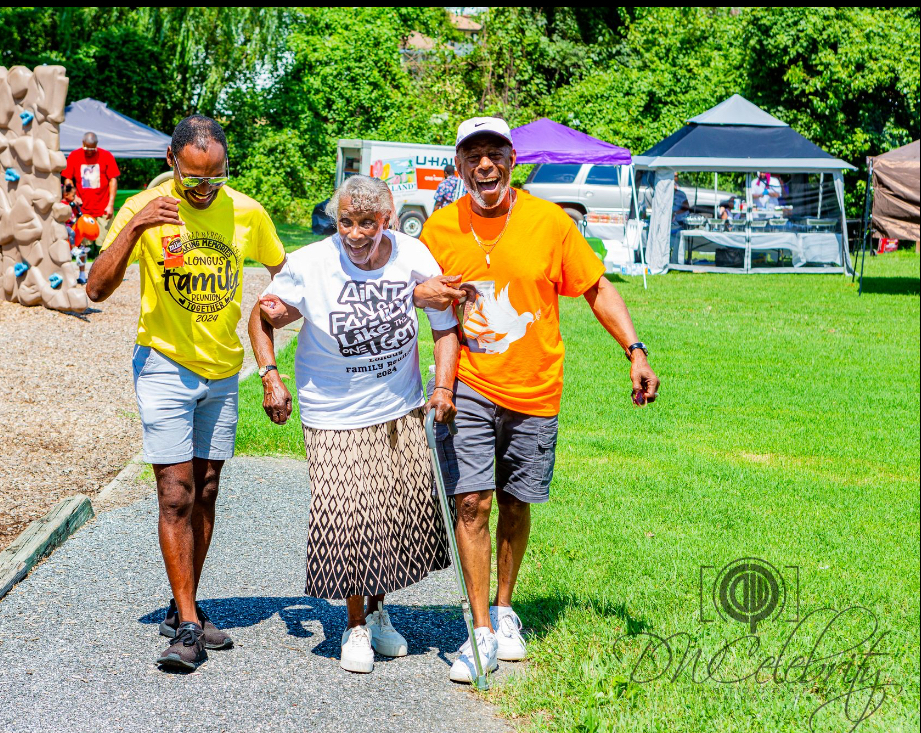 Elderly African American woman being escorted by 2 African American men along a path outside in a park