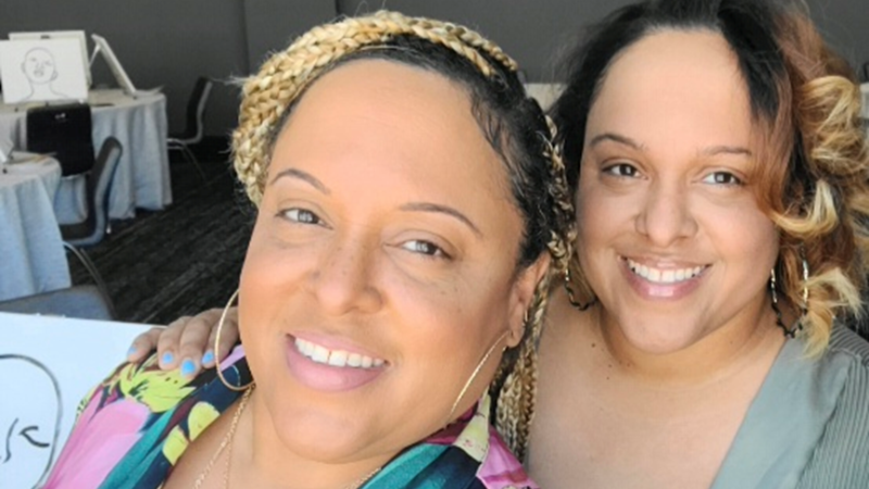 Two smiling twin sisters pose closely together indoors, one with blonde braided hair and a vibrant floral shirt, the other with curly dark hair with blonde tips and a muted green top, creating a warm, friendly atmosphere.